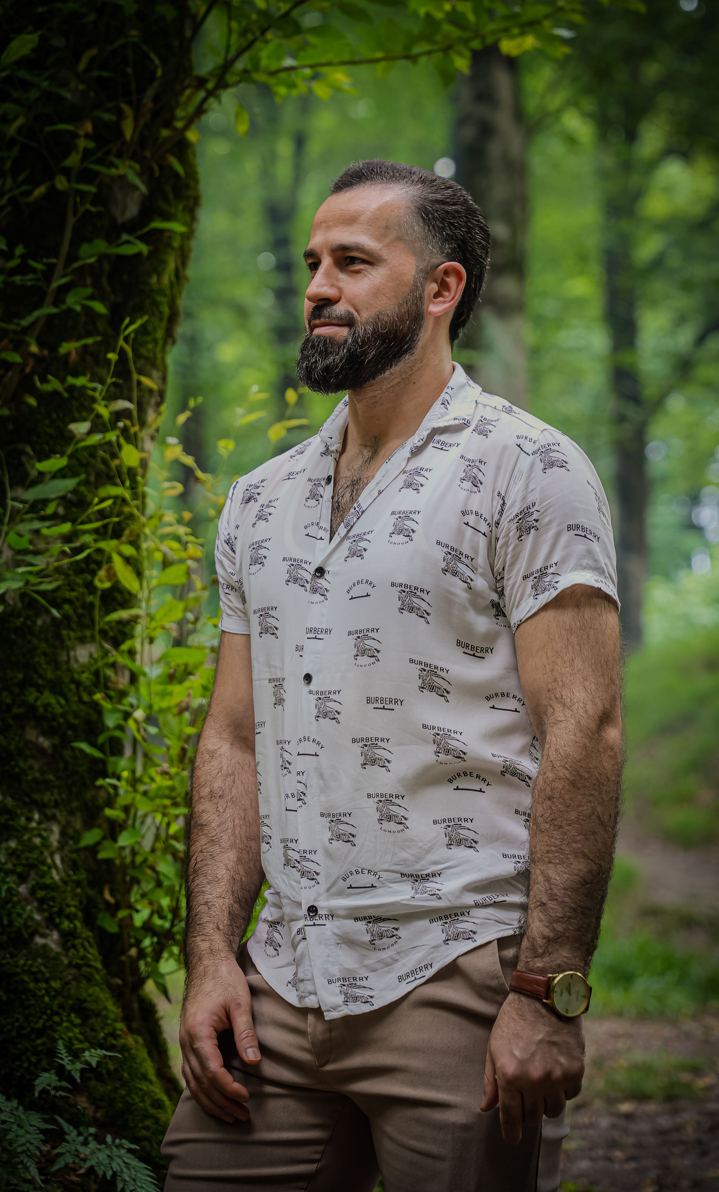 Bearded man standing in a lush Iranian forest, wearing a casual shirt.