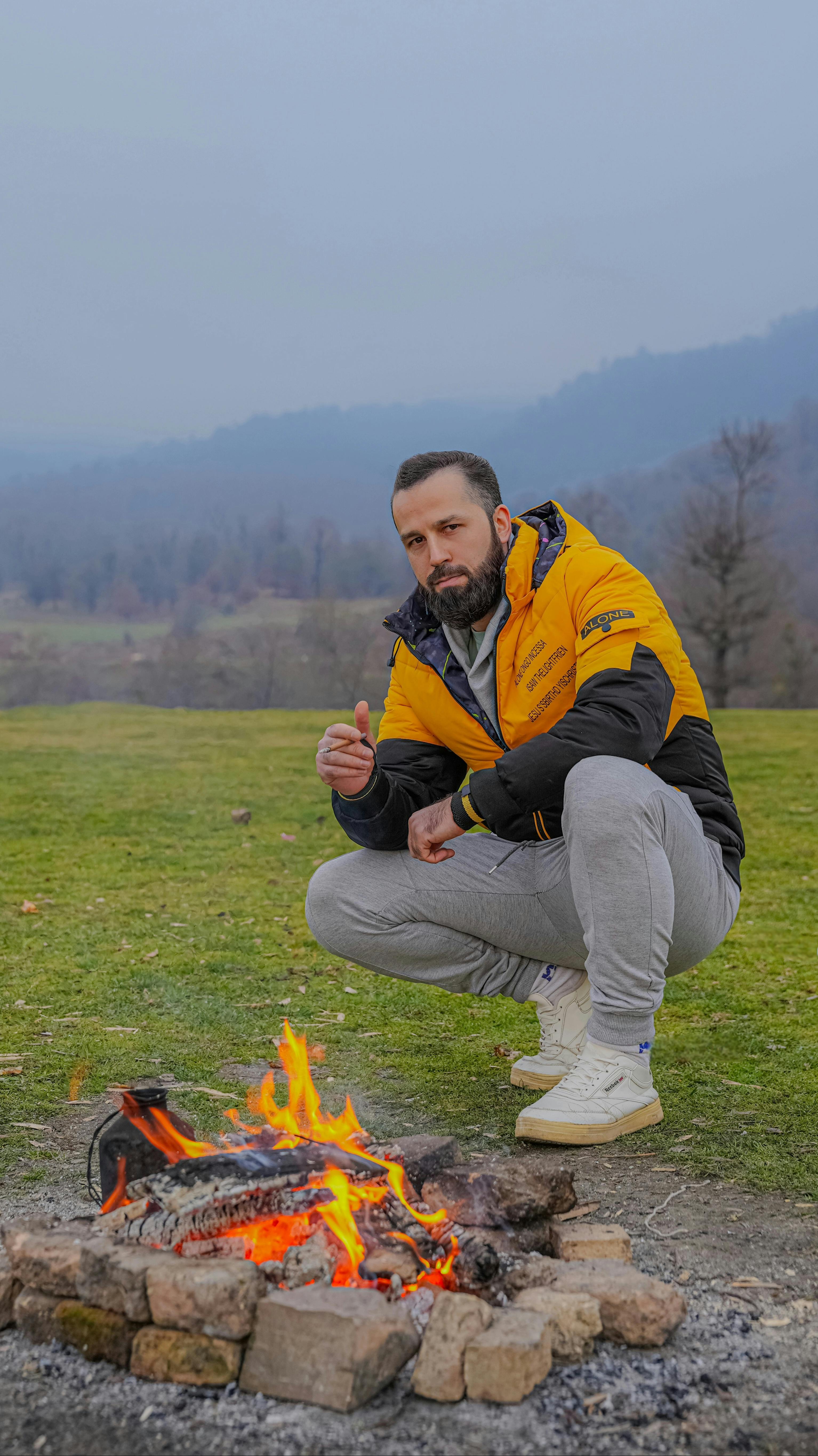 A man crouches by a vibrant campfire in a scenic outdoor setting in Iran, exuding a sense of adventure and warmth.