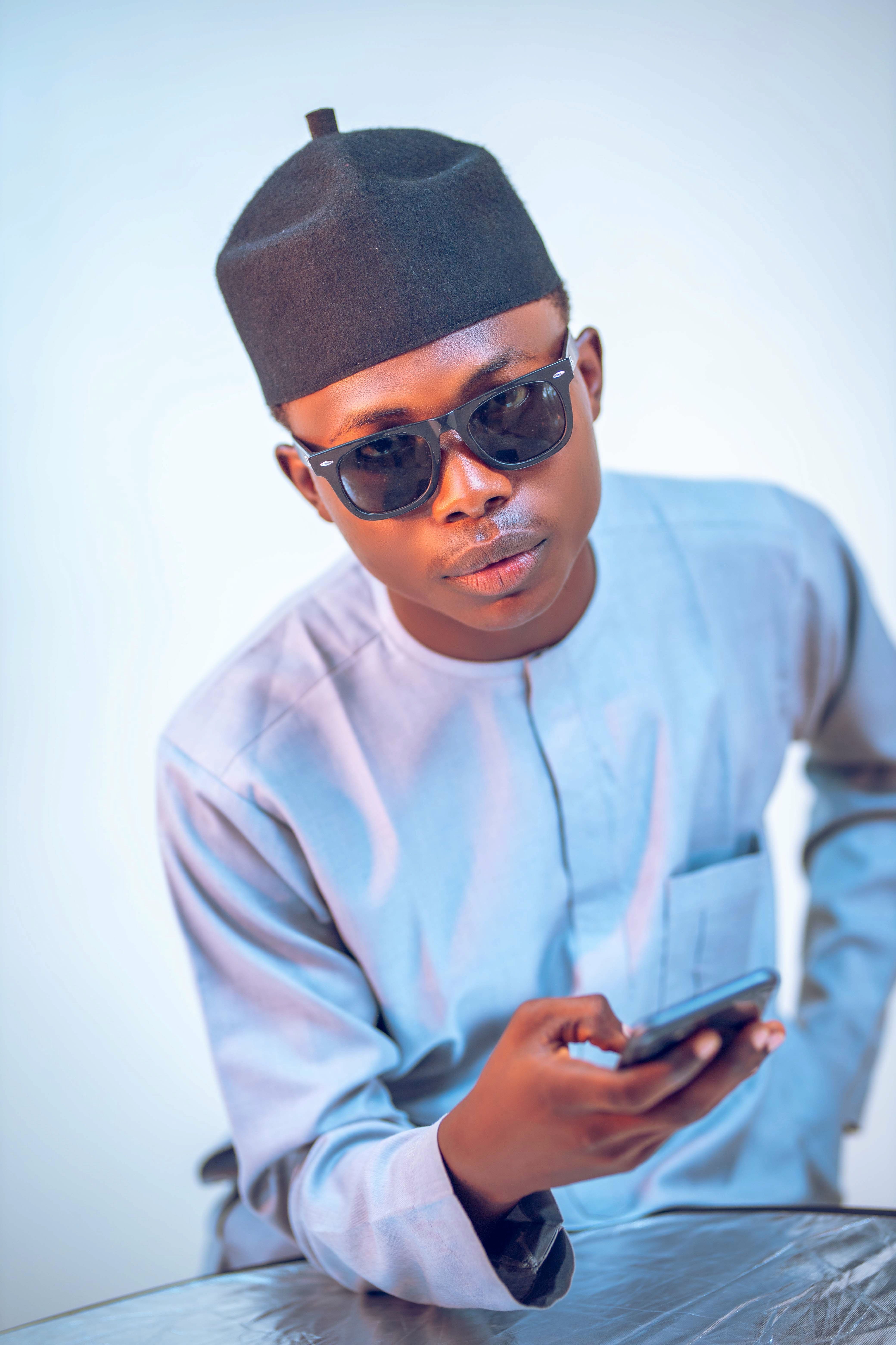 Young man wearing sunglasses and traditional attire, holding a smartphone indoors.