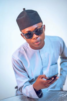 Young man wearing sunglasses and traditional attire, holding a smartphone indoors.