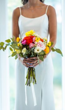 A bride in a white dress holding a vibrant bouquet of mixed flowers indoors.