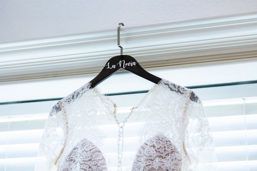 Elegant lace bridal gown with intricate details hanging on a personalized wooden hanger indoors.