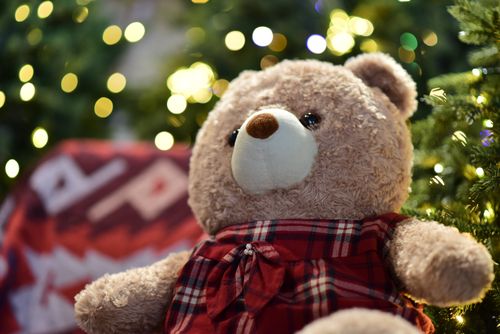 Free Cute teddy bear in red plaid outfit surrounded by holiday lights and decor, creating a warm festive atmosphere. Stock Photo