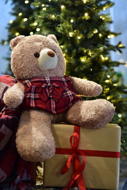 Free Cute teddy bear in plaid attire sits on gift box under a Christmas tree with lights. Stock Photo