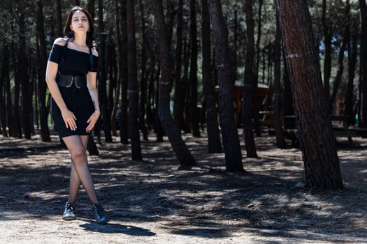 Young woman walking in a sunlit forest with tall trees around, wearing a fashionable black outfit.