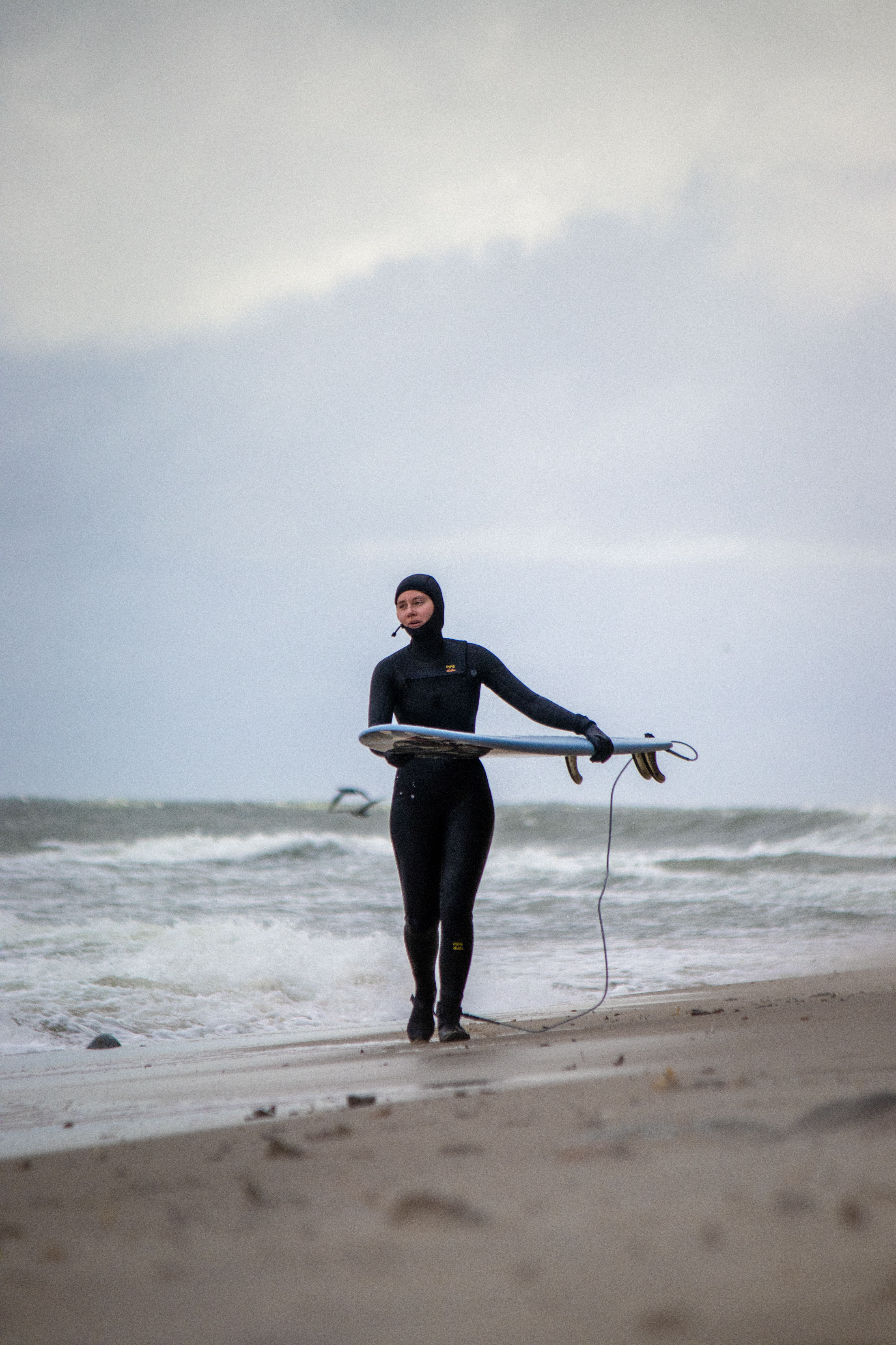 A woman in a wetsuit walks along the Baltic Sea coast with a surfboard, showcasing determination and adventure.