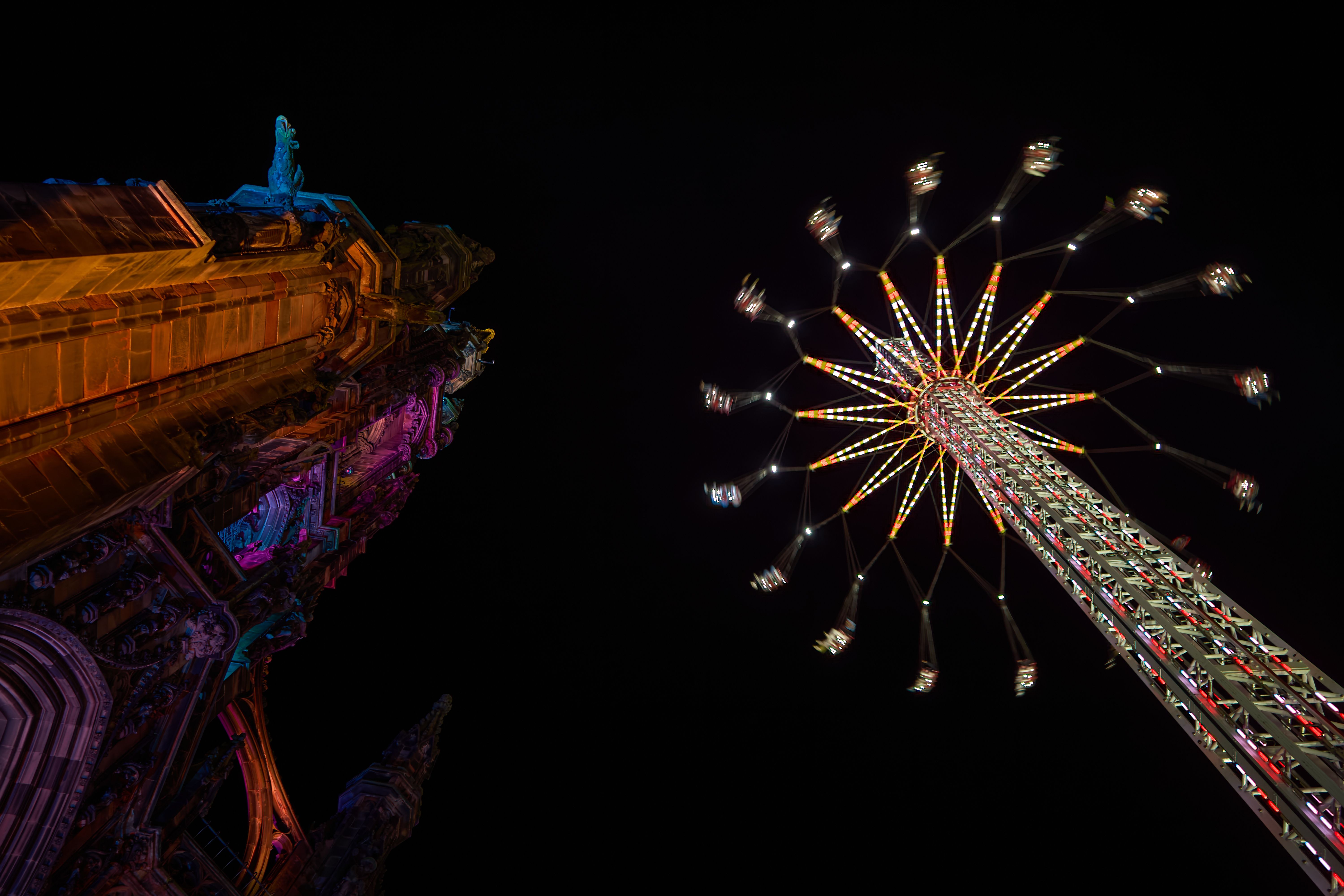 A tall Gothic stone spire lit in orange, pink and blue beside a lit swing ride spinning against a black sky