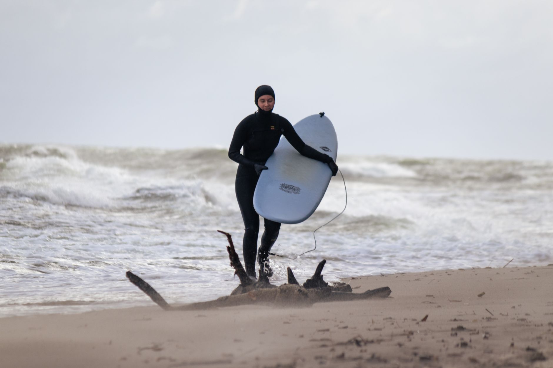 Un surfista en un traje de neopreno camina por una playa fría y ventosa llevando una tabla de surf con olas rompiendo cerca.