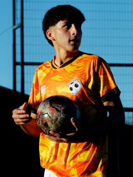 Young soccer player holding a ball on an outdoor field during the day. Bright sports jersey under blue sky.
