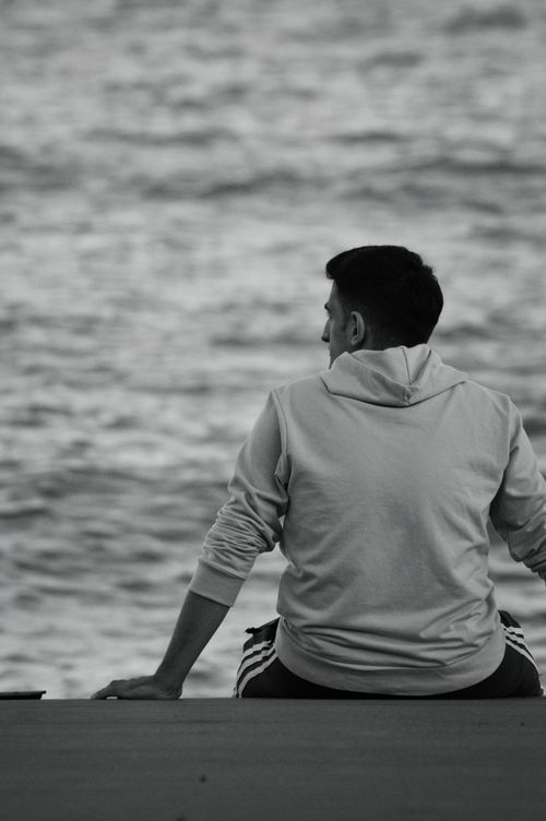 Free A person in a hoodie sits by the ocean, reflecting in a calm and serene setting. Stock Photo