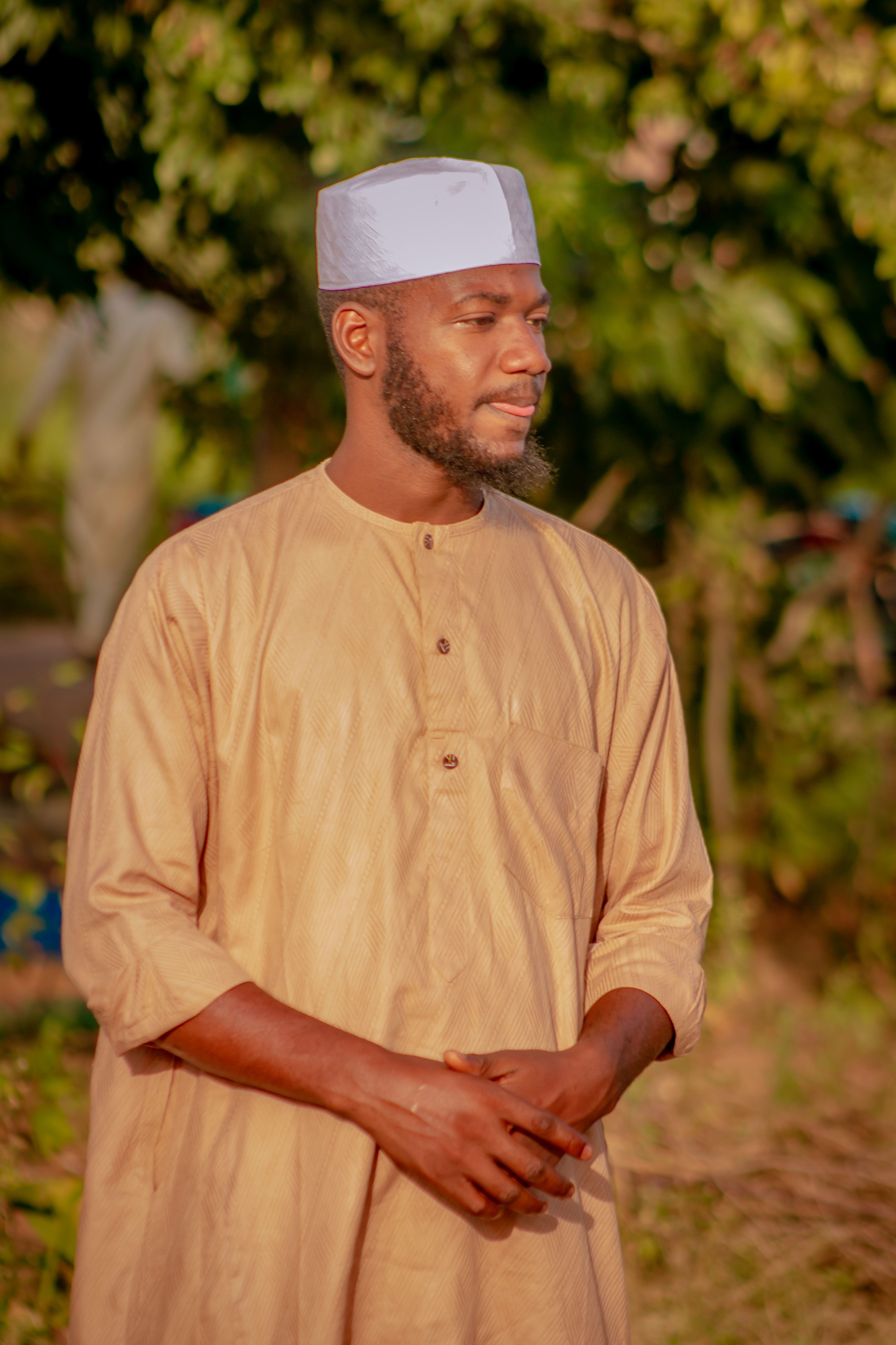 A man in traditional attire stands outdoors, surrounded by greenery, reflecting a cultural lifestyle.