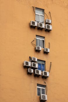 Yellow building facade with windows and numerous air conditioning units attached.