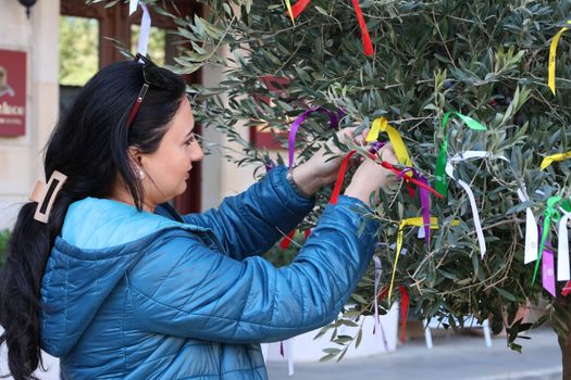 An adult woman ties colorful ribbons onto a tree branch during a daytime outdoor activity.