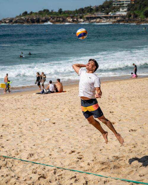 Free Energetic man leaps for a volleyball on a lively beach, surrounded by people enjoying the sun and sea waves. Stock Photo