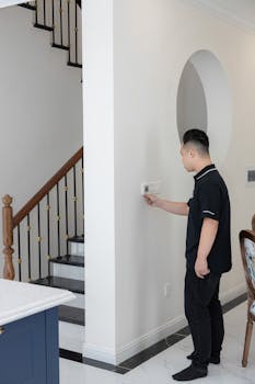 A young man adjusts the thermostat on a wall in a modern indoor setting with a staircase.