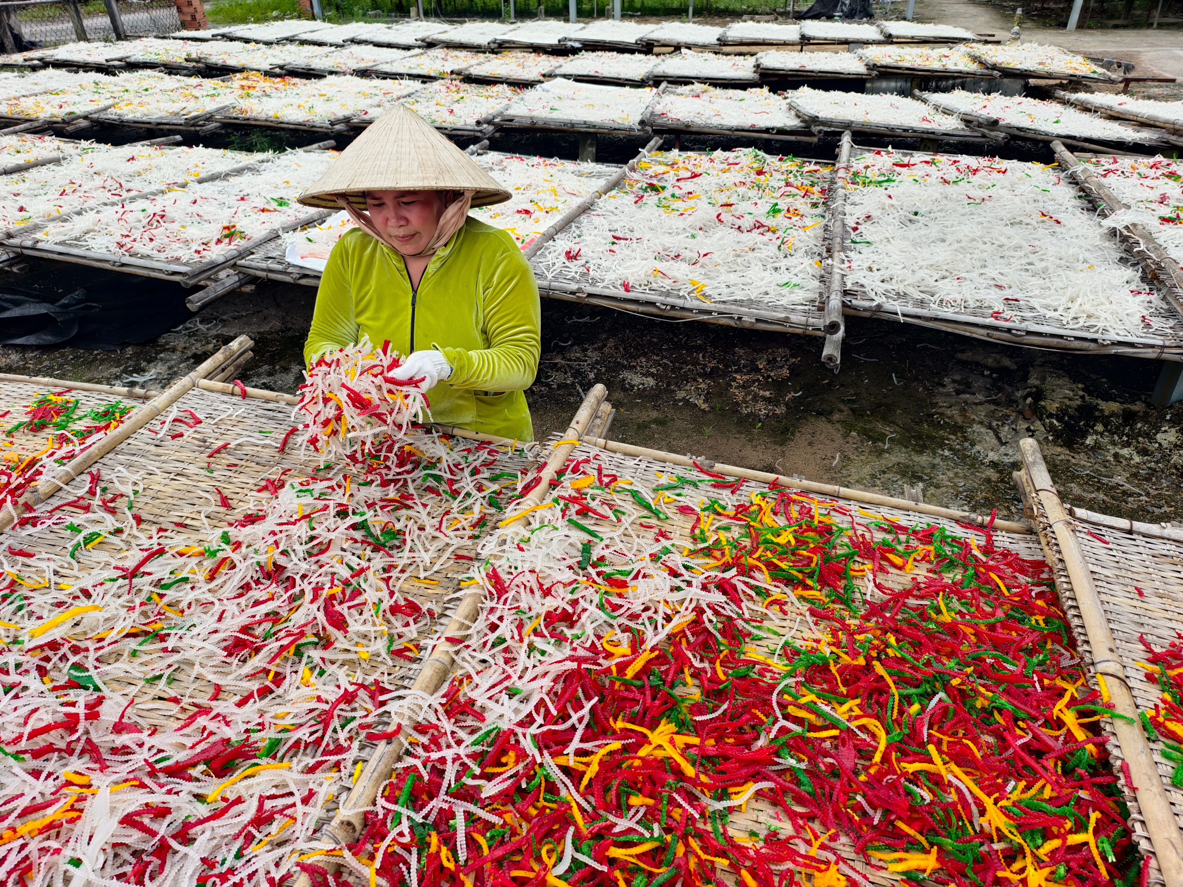 Person arranging colorful dried vegetables on bamboo racks in an outdoor setting.