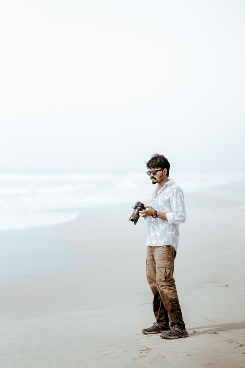 Free Man holding a camera on a tranquil beach, capturing serene ocean views. Stock Photo