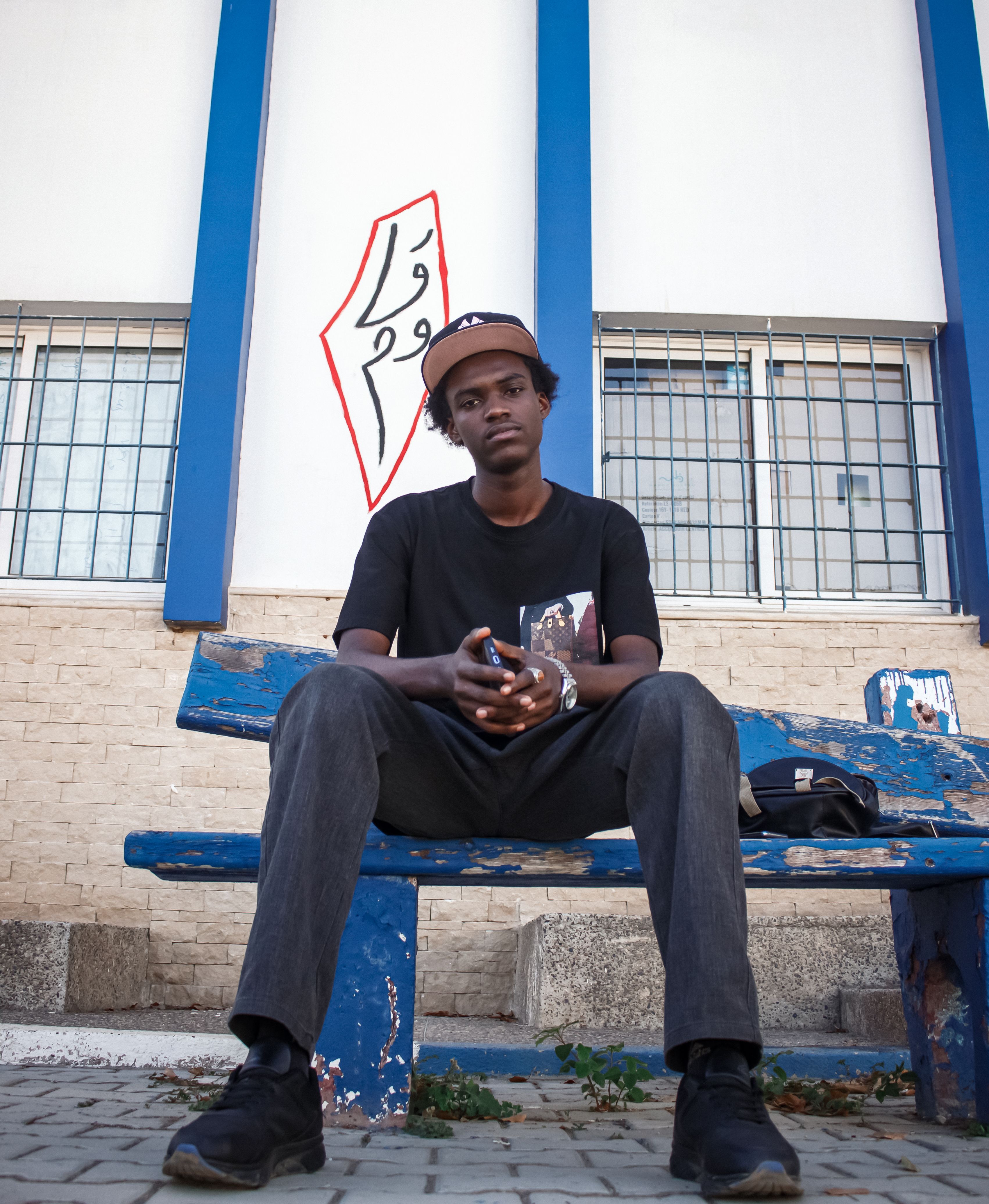 A person sits on a worn blue bench against a graffiti-adorned wall.