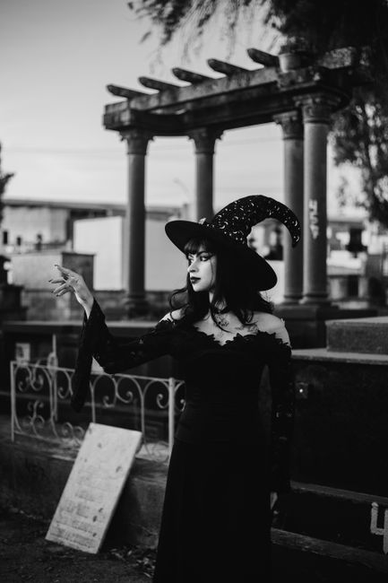 A gothic witch stands in a cemetery wearing a black hat and dress, evoking a mystical atmosphere.