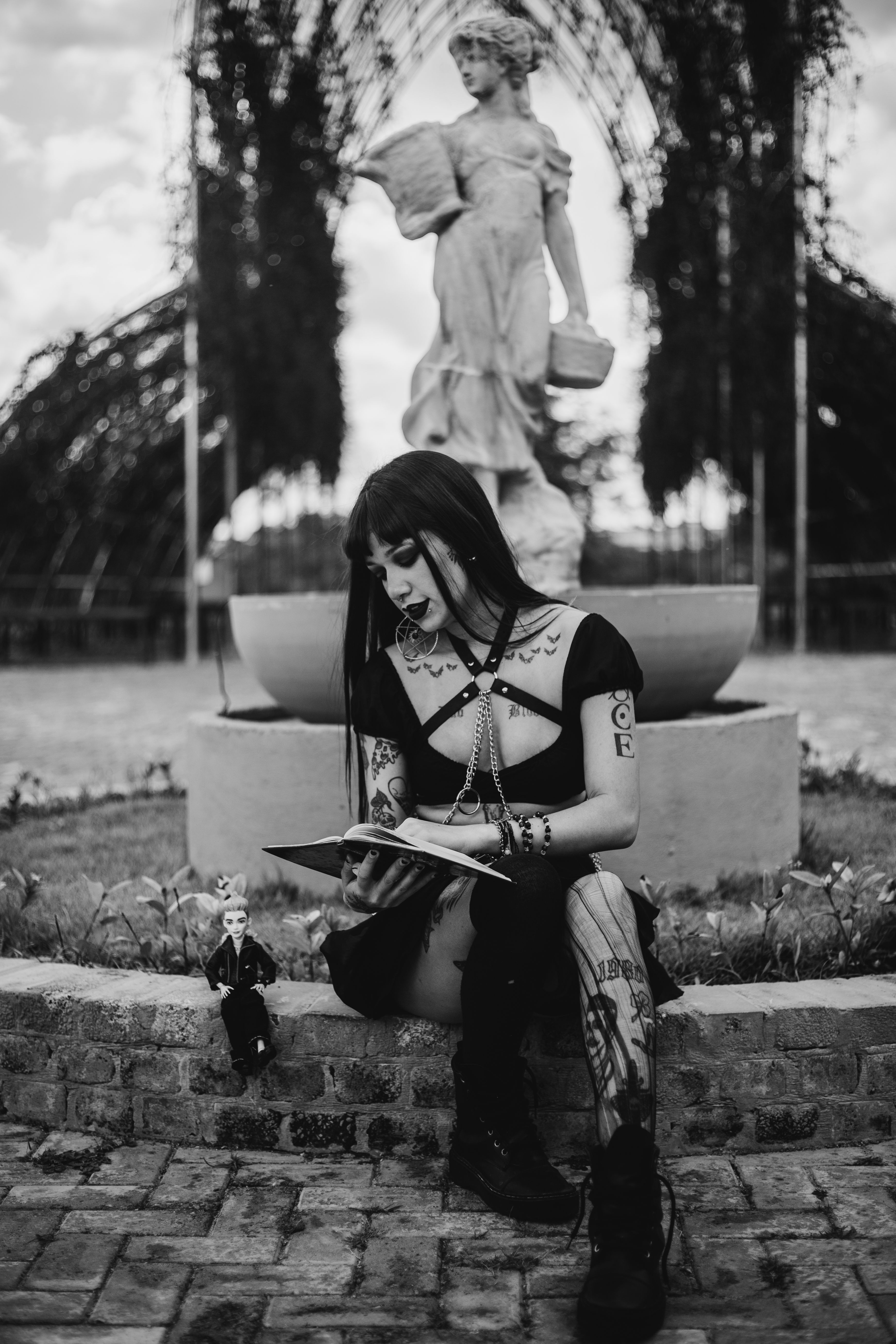 Gothic woman with tattoos reading a book near a classic statue outdoors.