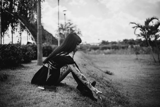 A contemplative tattooed woman sitting on grass in a black and white outdoor scene.