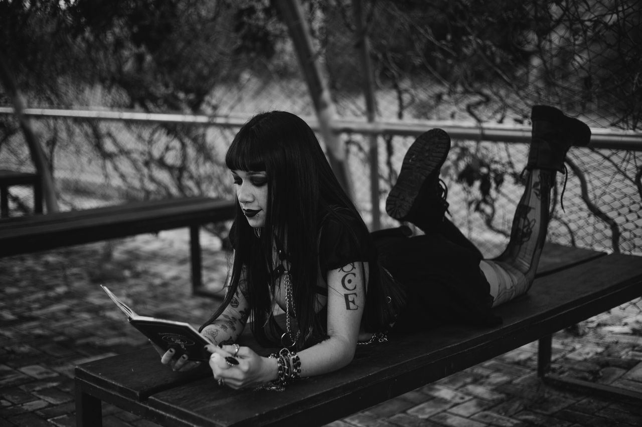 a gothic girl with tattoos lies on a bench reading in a moody black an