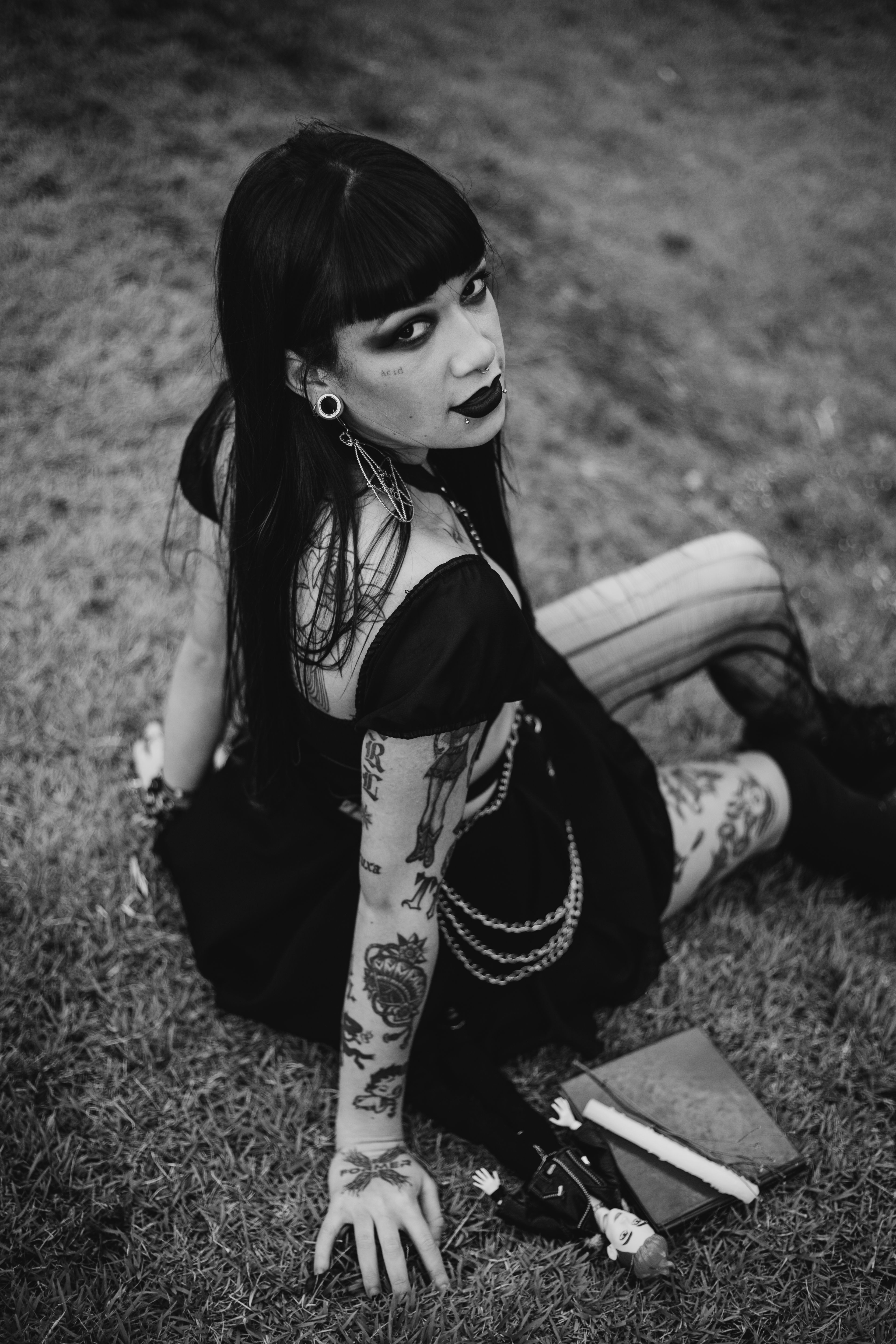 A black and white portrait of a woman with tattoos and gothic style, sitting outdoors on grass.