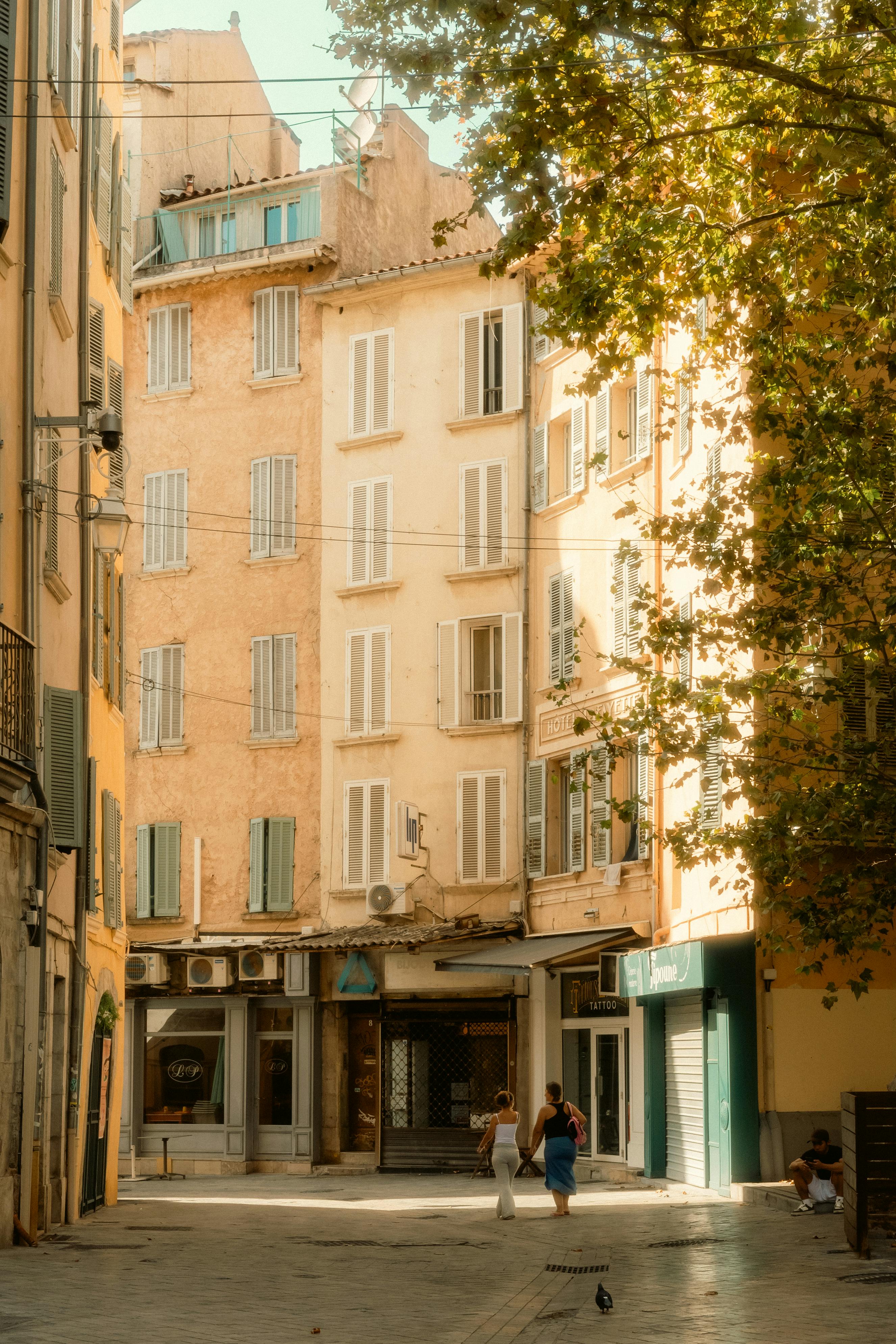 Golden light bathes a quaint European alleyway with pedestrians and shuttered windows.