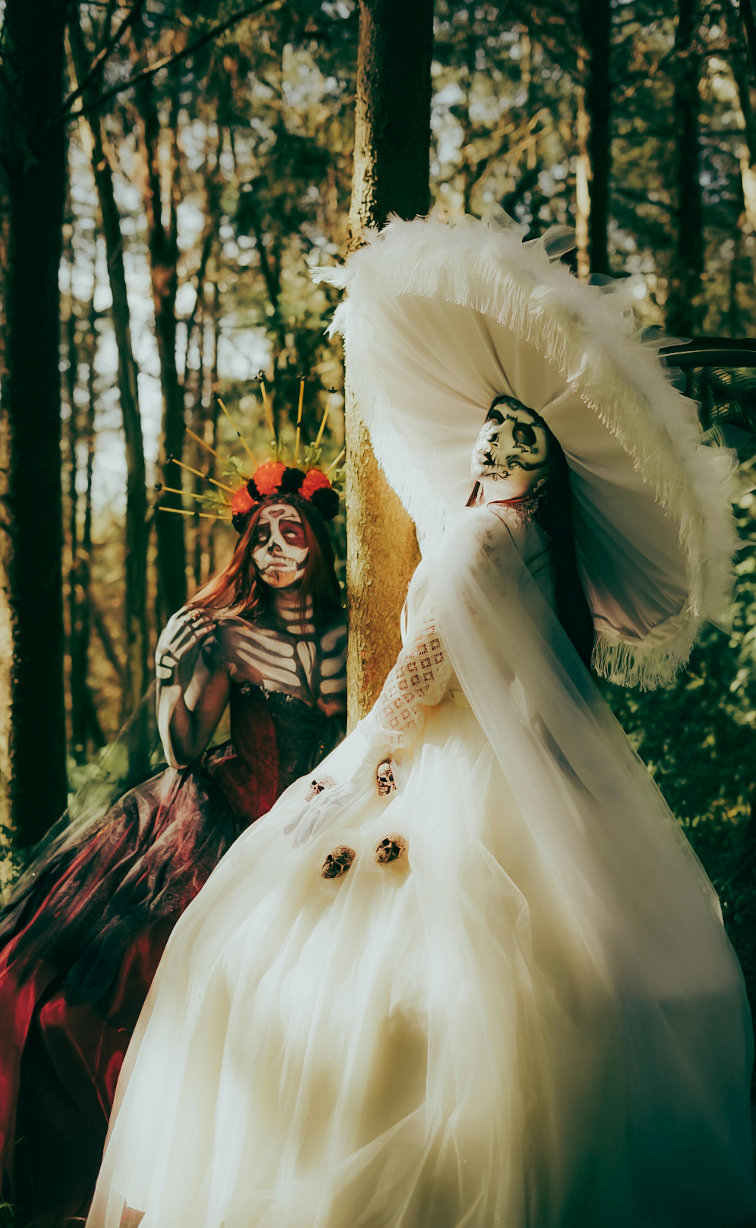 Two individuals in intricate Day of the Dead costumes pose in a sunlit forest setting.