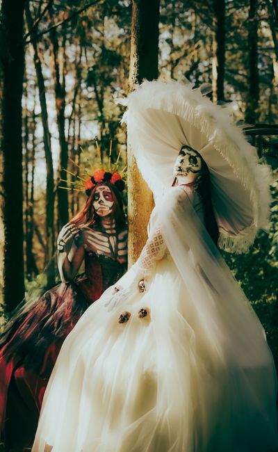 Two individuals in intricate Day of the Dead costumes pose in a sunlit forest setting.