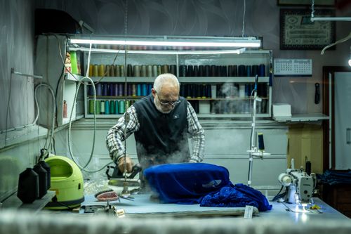 Free Elderly tailor ironing blue fabric in a traditional sewing workshop surrounded by sewing tools. Stock Photo