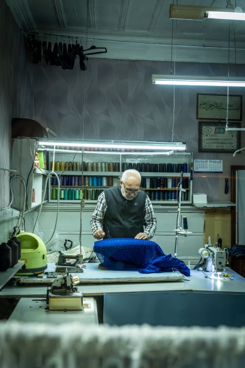 Free An experienced tailor crafting with fabric on a sewing table, surrounded by thread spools in a well-lit workshop. Stock Photo
