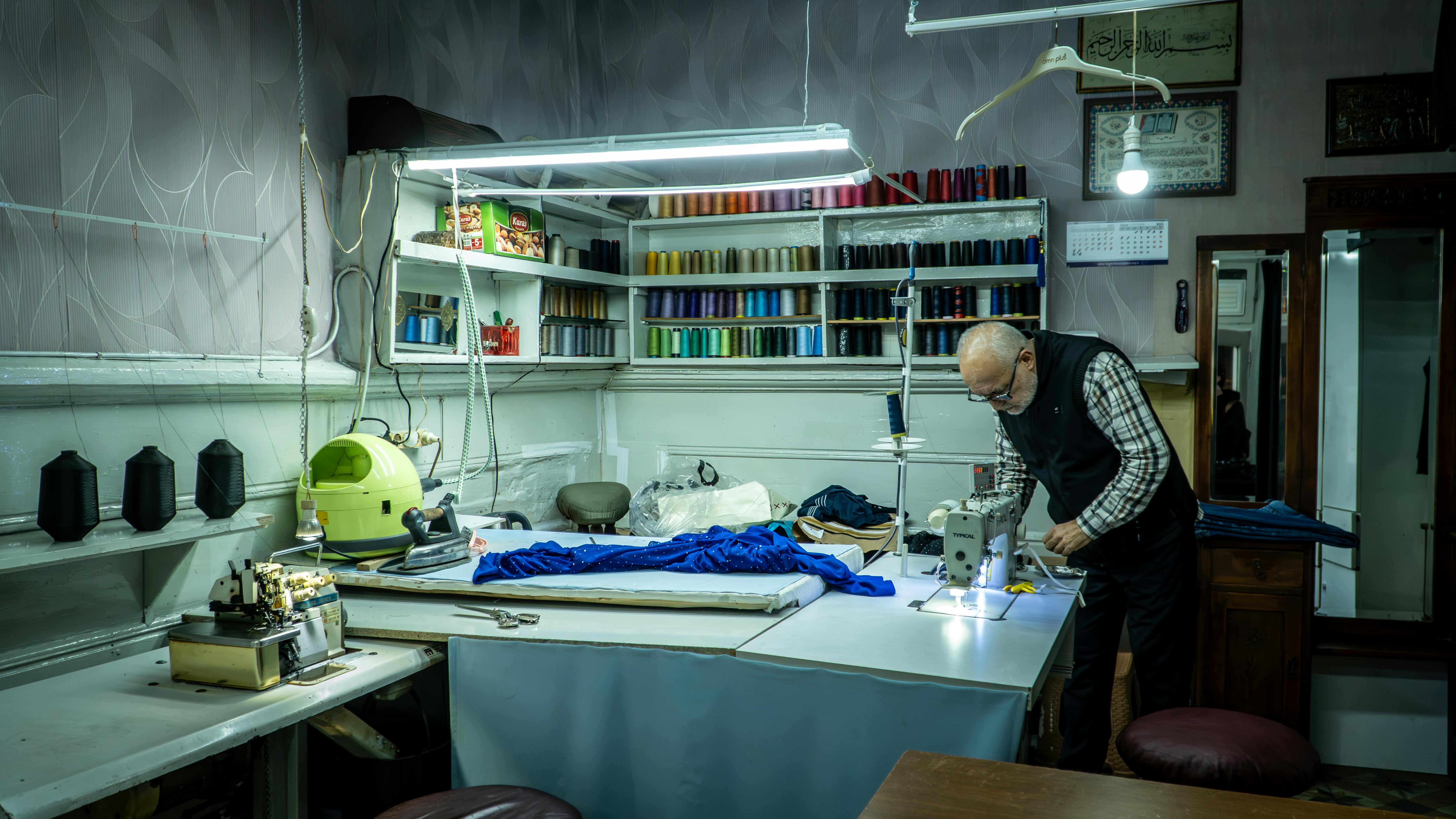 An elderly man sewing in a workshop with colorful threads and sewing machines, showcasing craftsmanship.