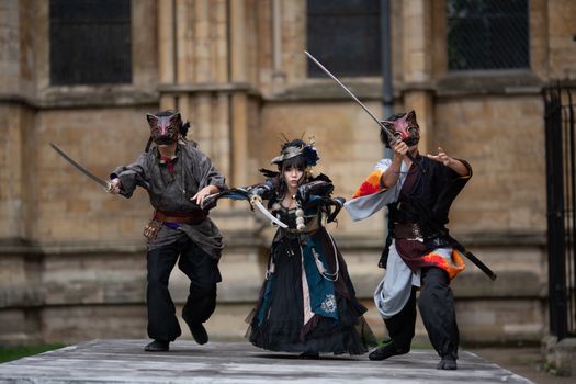 Three people in intricate fantasy samurai costumes pose dramatically outdoors, wielding swords.