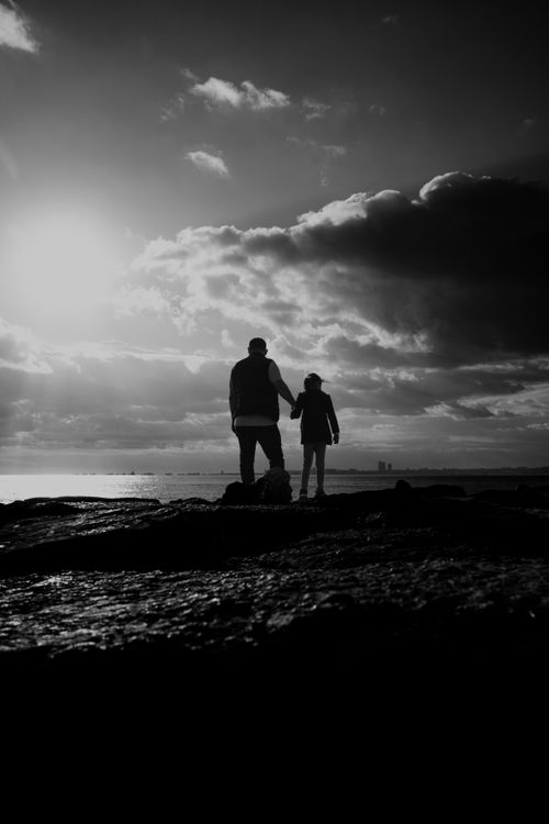 Free A heartwarming black and white silhouette of a father and child holding hands on a rocky shore during sunset. Stock Photo