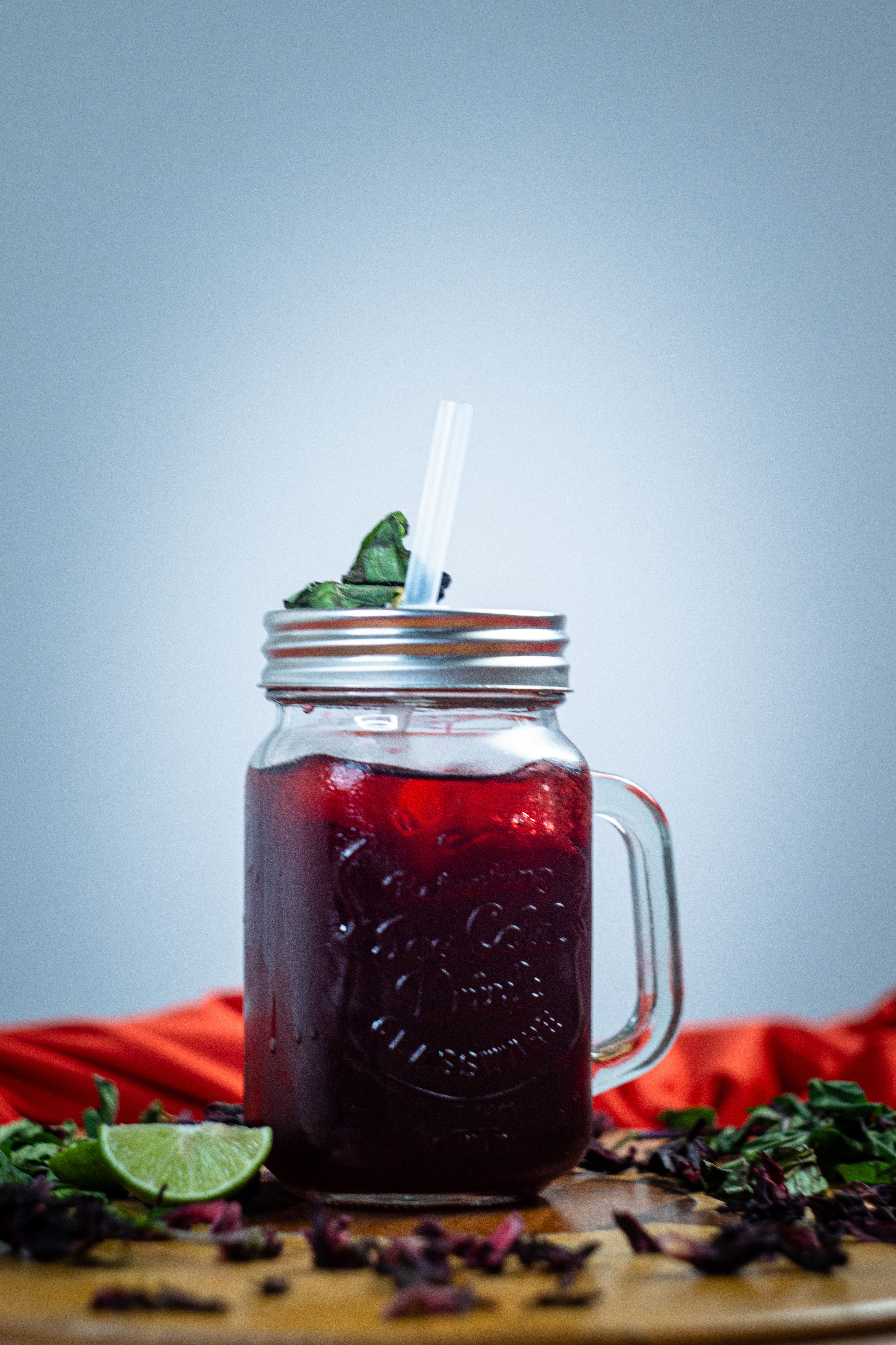 A glass pitcher of deep red agua de jamaica filled with ice, with a glass poured alongside dried hibiscus flowers and lime wedges on the table.