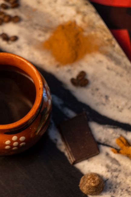 A steaming mug of frothy Mexican hot chocolate in a traditional clay jarrito, topped with foam and a cinnamon stick, next to broken pieces of a Mexican chocolate tablet.