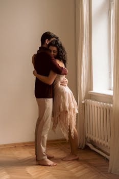 A couple shares a tender embrace indoors near a sunlit window.
