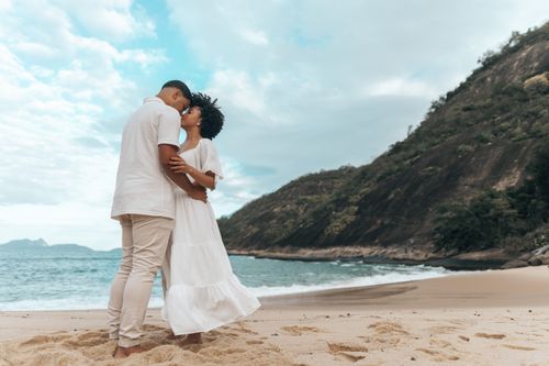 Free A couple embracing on a scenic beach with hills in the background under a cloudy sky. Stock Photo