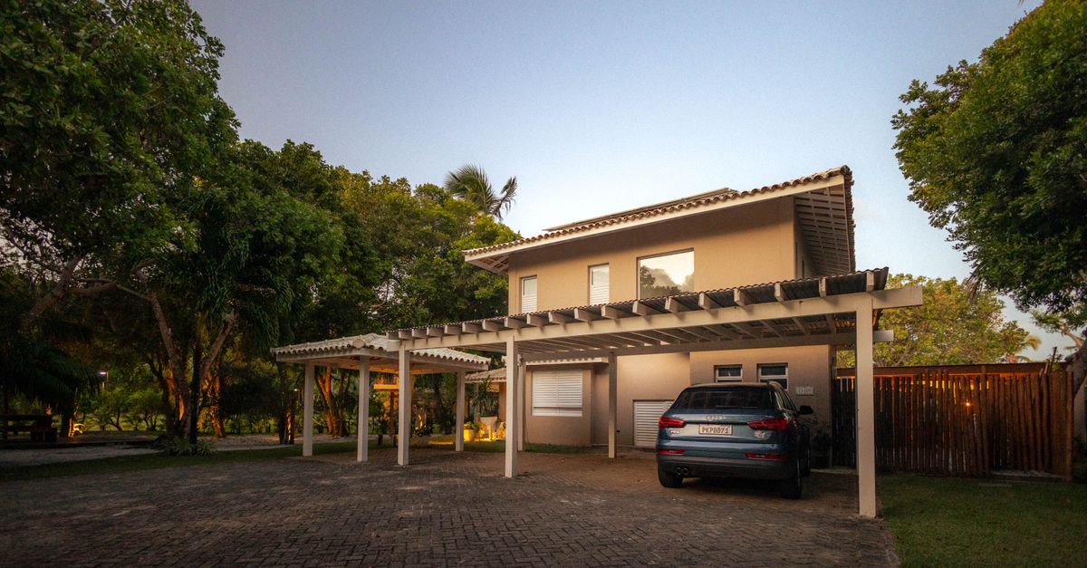 A modern tropical house with a pergola and driveway at twilight, surrounded by lush greenery.