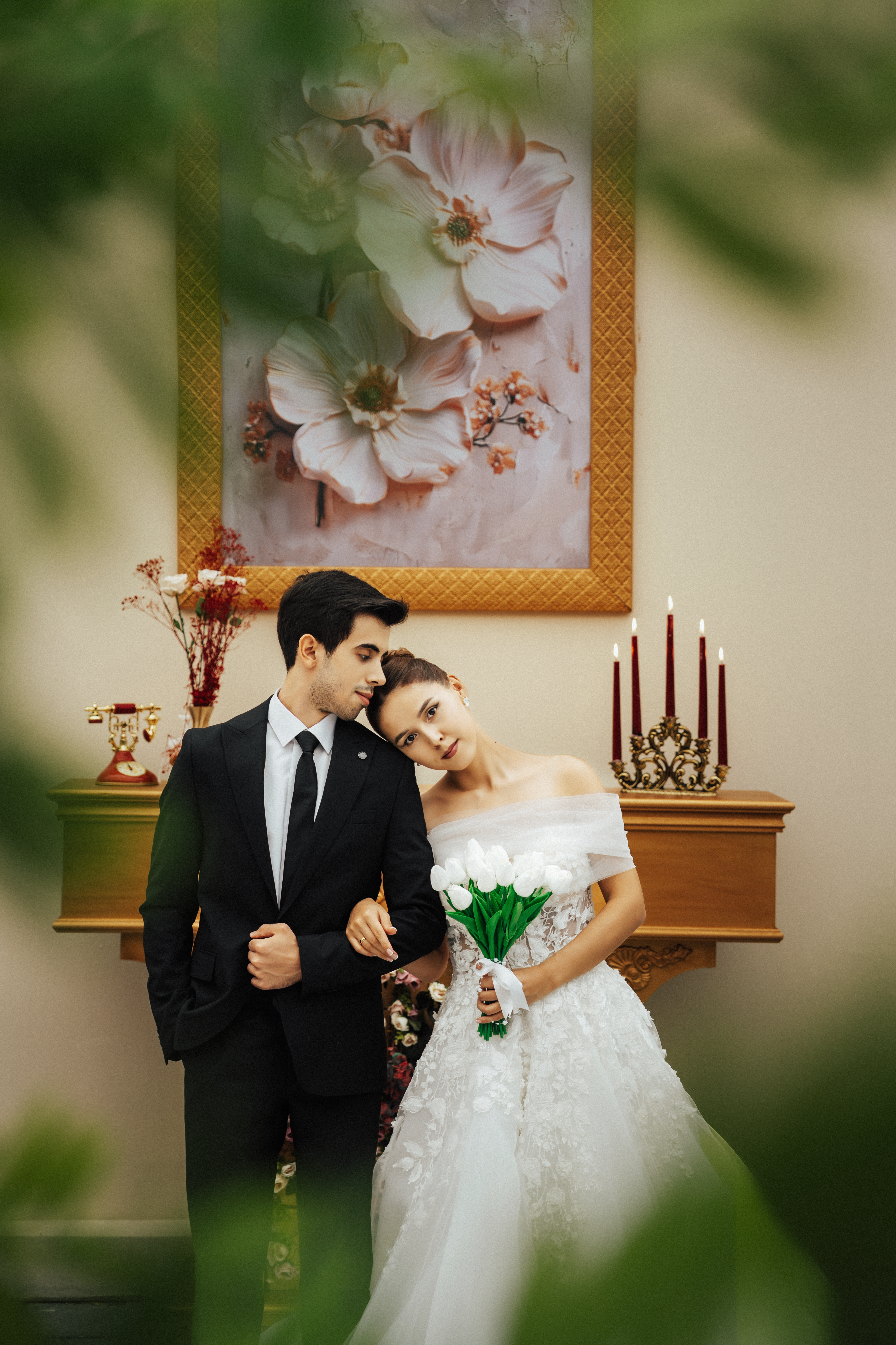 An elegant wedding couple poses indoors with floral art and candlesticks, creating a romantic atmosphere.