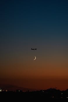 Silhouette of an airplane flying above a crescent moon against a vivid sunset sky.