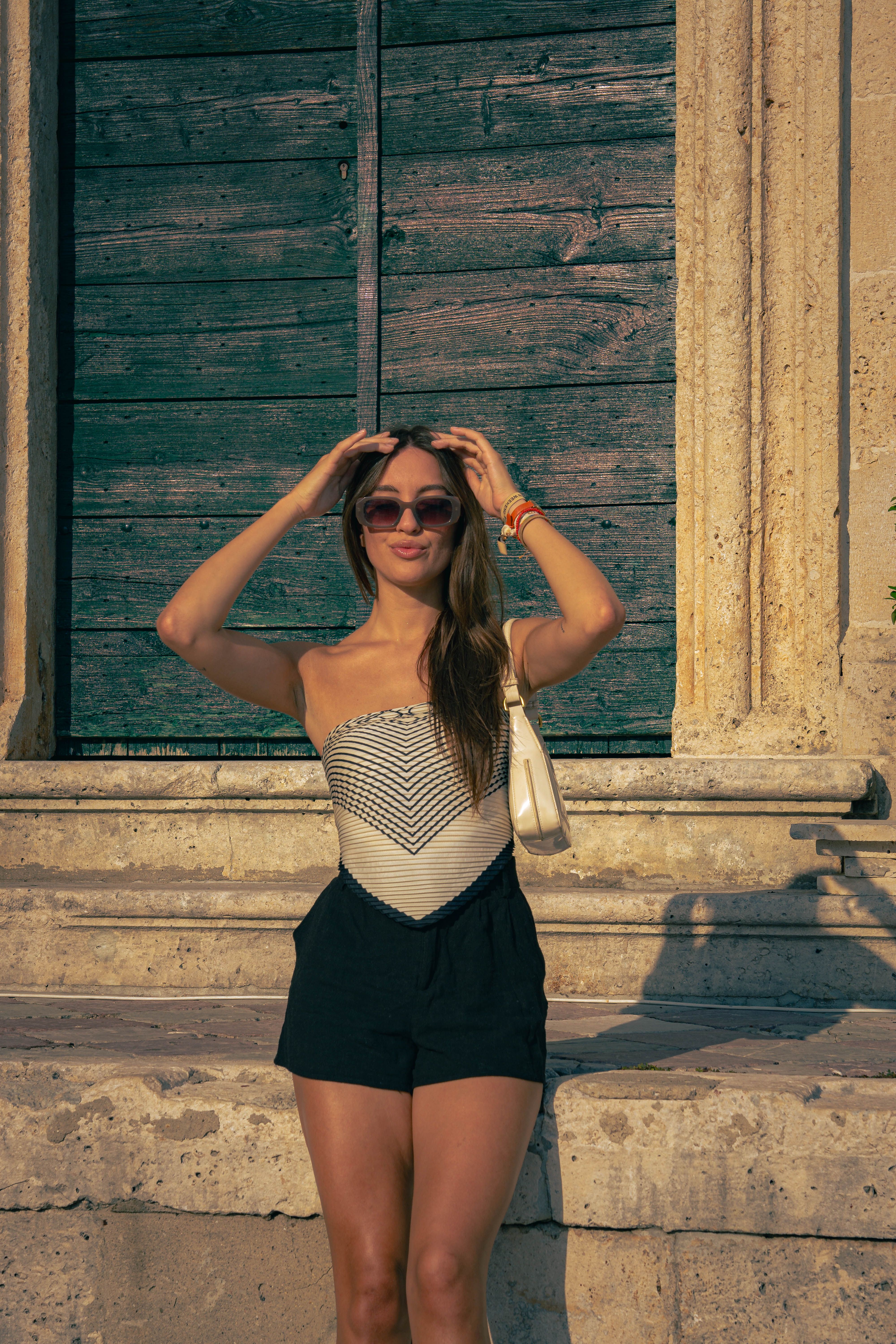 Femme élégante portant des lunettes de soleil, posant près d'une porte en bois sous un soleil radieux. 