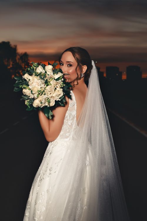 Free A beautiful bride in a detailed white gown holding a bouquet during a sunset, exuding elegance and joy. Stock Photo