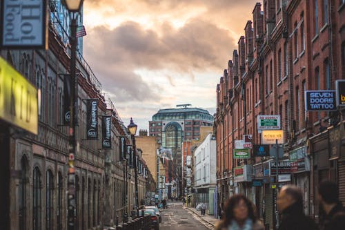 Person on Road Between Brown Walled Building during Daytime Under Cumulus Clouds