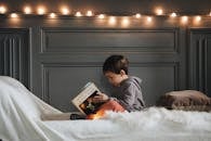 一个男孩在舒适的卧室里看书，卧室里挂着温暖的串灯，铺着柔软的纹理
