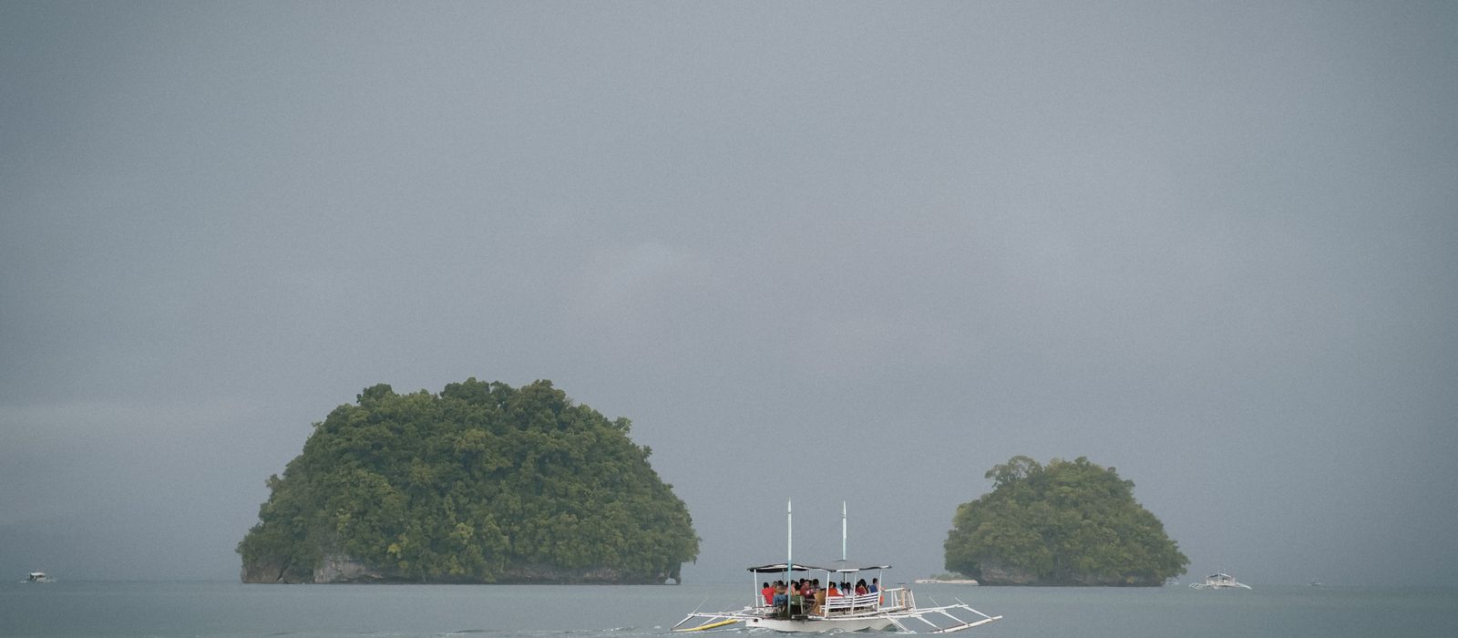 A boat navigates between lush tropical islands on a misty day, evoking serenity.