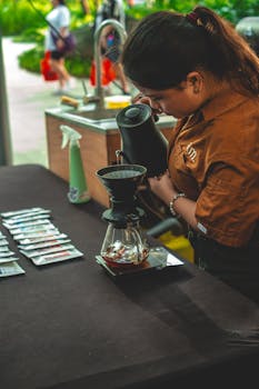 A barista brews coffee using a pour-over method at an outdoor park event.