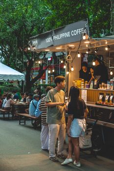 People enjoying coffee at an outdoor stall in a lively city park setting.