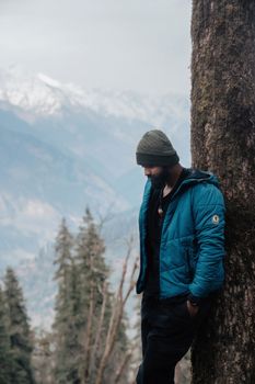 Man standing by a tree, gazing at snow-capped mountains in a tranquil winter setting.